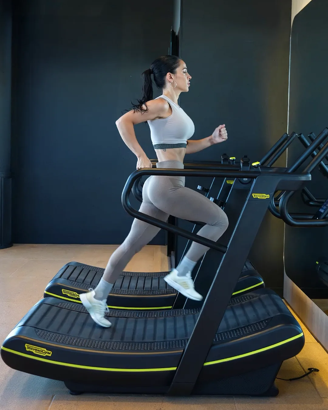 A member at pace on the curved Technogym treadmill in the dark-walled cardio room